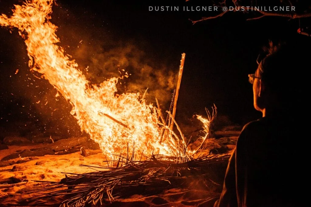 Person watching a large bonfire on a sandy beach, demonstrating how to cook in the wild.