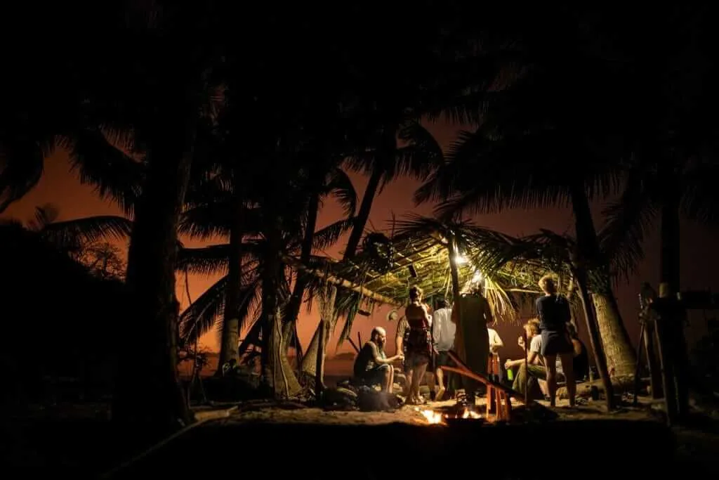Survival on a desert island at night with a small makeshift shelter and a group of people around a campfire.