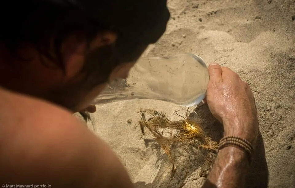 A man demonstrates how to survive on a desert island by holding a water bottle in the sand.
