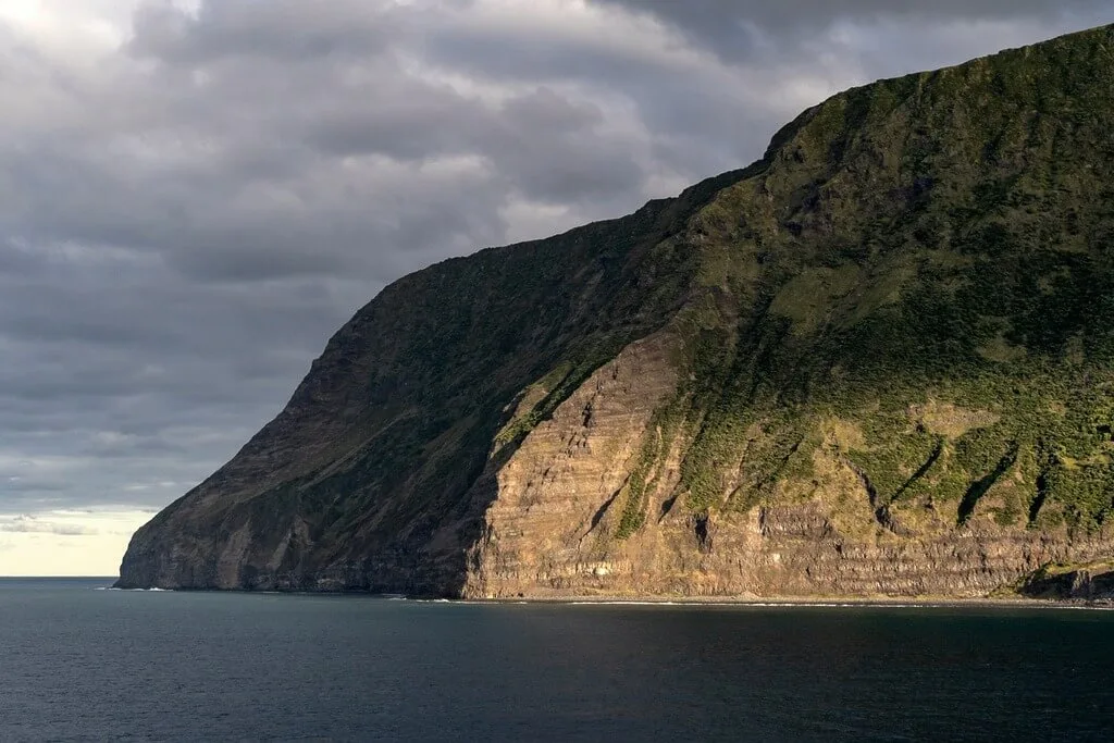 Remote island coastline with a steep, green-covered cliff face meeting the dark ocean under a cloudy sky.