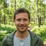 A smiling man in a forest setting, wearing casual outdoor clothing, surrounded by tall trees and lush greenery, emphasizing wilderness exploration and survival skills.