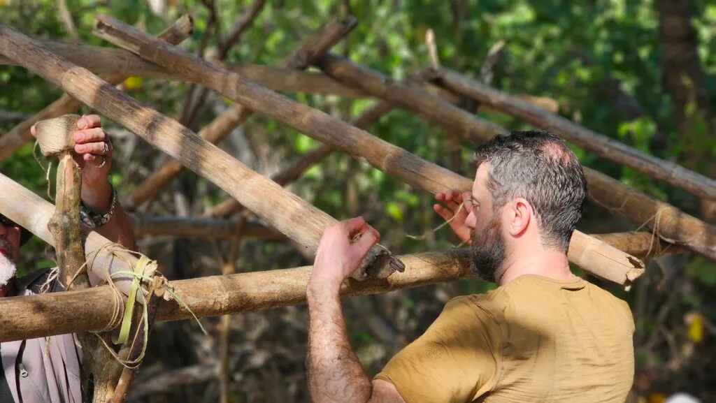 Two men collaborate outdoors, assembling a wooden structure from thick branches and tying them with natural fibers—a great team building activity surrounded by trees and greenery.
