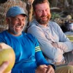 Two men sit by a palm tree at the beach, smiling and laughing together during a team building retreat. One wears a blue shirt and cap, holding a coconut, while the other wears a light gray hoodie. The background includes sand and palm trees.
