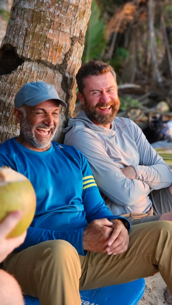 Two men laughing during team building, sitting by a tree with a coconut.