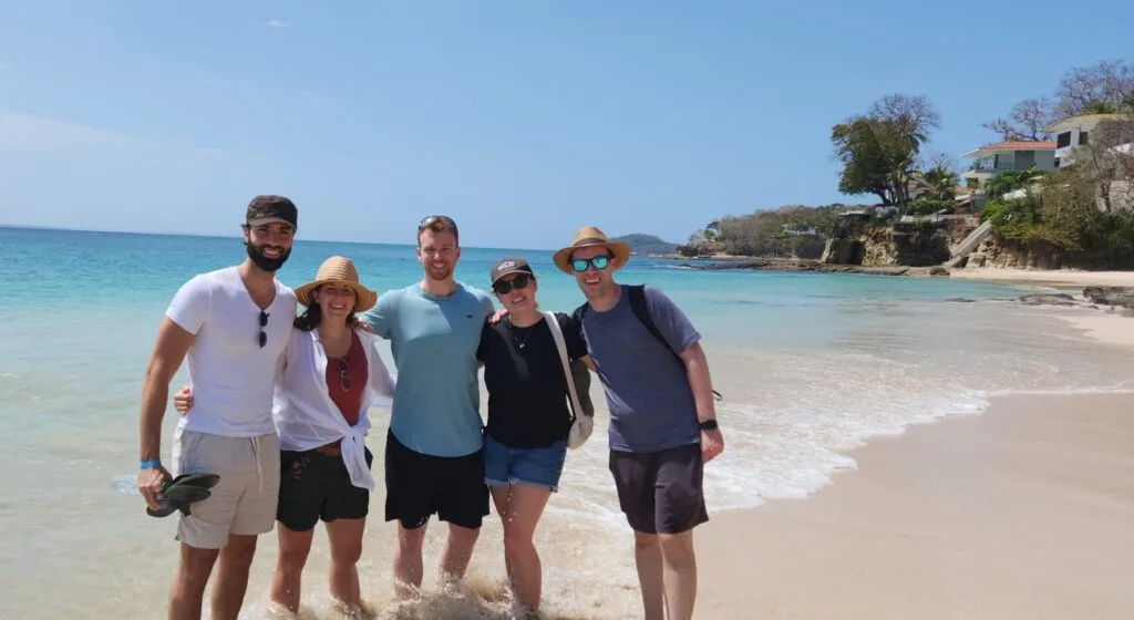 Five friends, enjoying a team building day, stand smiling together on a sandy beach with clear blue water and sunny skies. Wearing casual summer clothes, hats, and sunglasses, some dip their feet in the water; houses and trees are in the background.