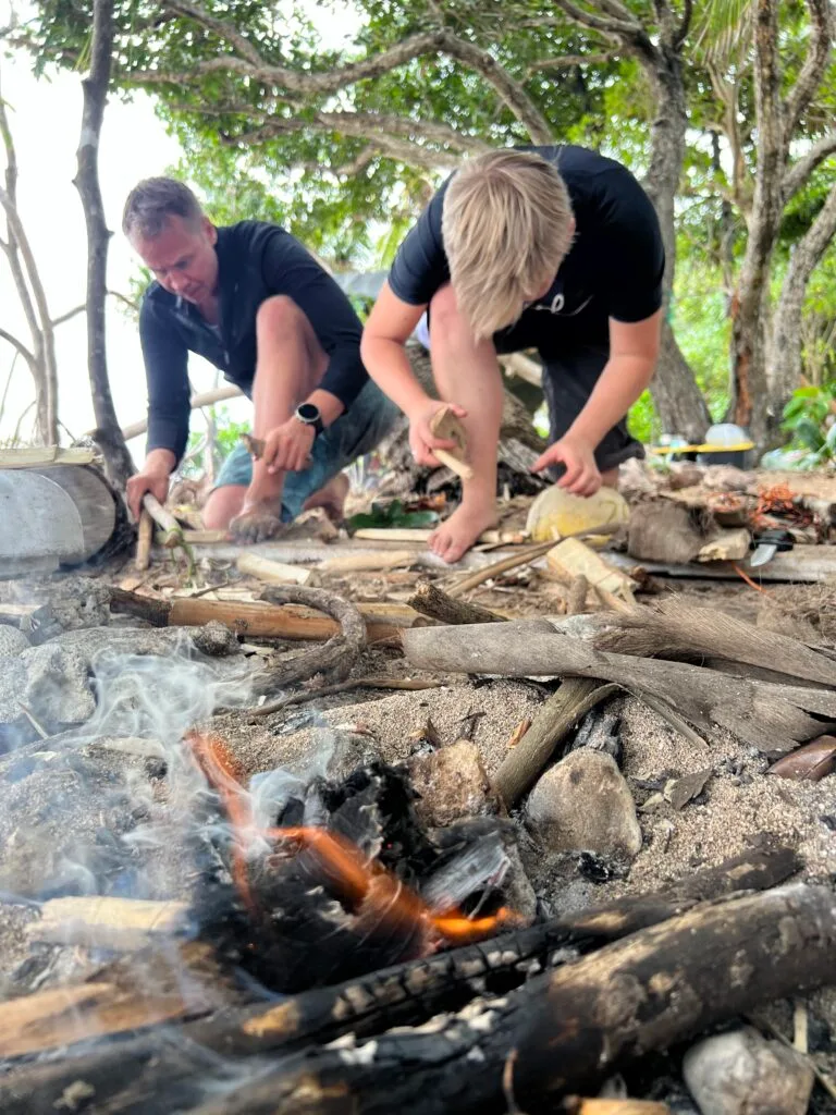 Two people crouch on a beach surrounded by driftwood, using tools to start a fire—an engaging team building activity. Smoke and small flames rise from burning wood, with trees and greenery in the background.