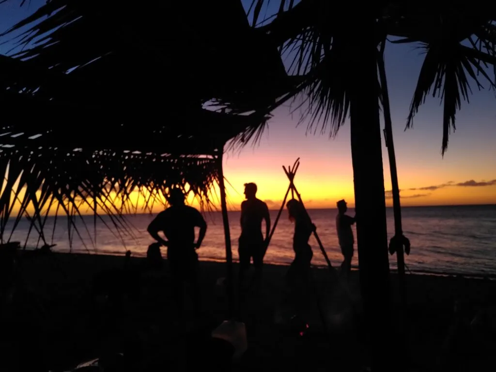 Silhouettes of four people stand under palm fronds on a beach at sunset, capturing a team building moment against the ocean and a colorful sky of yellow, orange, and purple hues in the background.
