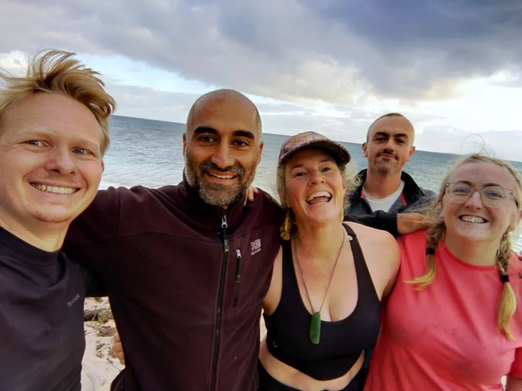 Five smiling adults stand close together on a beach with the ocean and cloudy sky in the background, appearing happy and relaxed as they pose for a group selfie during a team building retreat.
