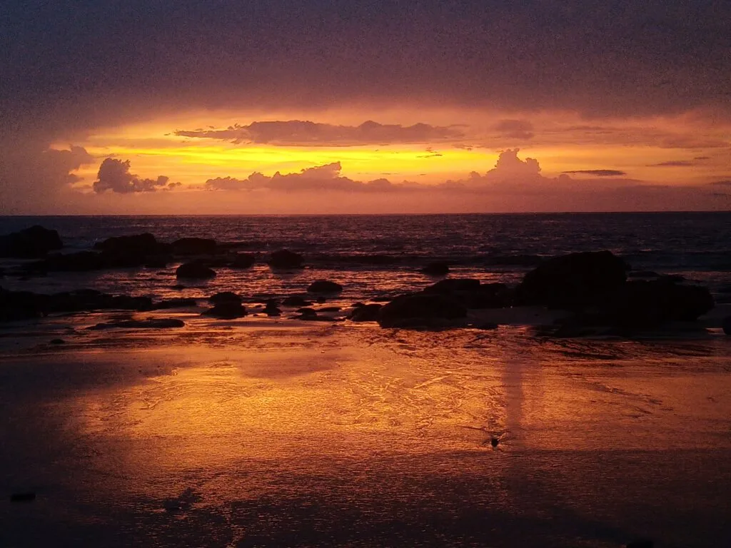 A dramatic sunset over the ocean, with orange and yellow light reflecting on wet sand and scattered rocks along the shore, creates a perfect setting for team building under a cloudy sky.