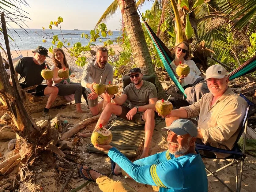 Six people sit on a sandy beach under palm trees, enjoying team building as they smile and hold coconuts with straws. The ocean glimmers in the background, and sunlight filters through the trees, creating a relaxed, tropical atmosphere.