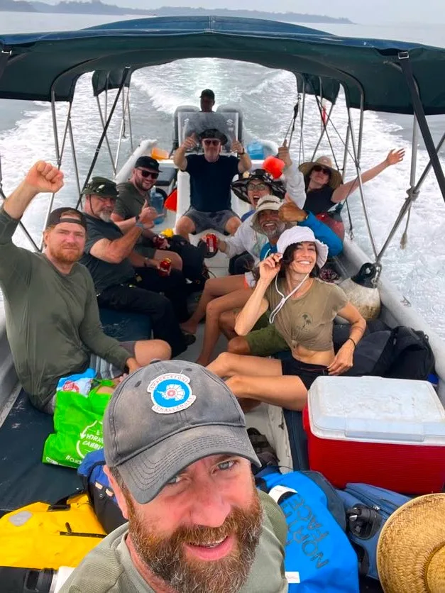 A group of people smile and pose for a selfie on a small motorboat filled with gear and coolers, enjoying a fun team building adventure under a canopy, with waves trailing behind them.