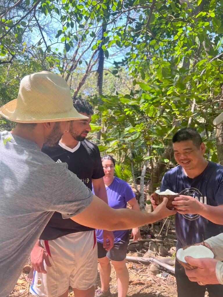 Four people stand together outdoors in a sunlit, leafy area, enjoying a team building activity. One man in a gray shirt and hat reaches out to receive a coconut shell from another smiling man, while two others look on in the background.