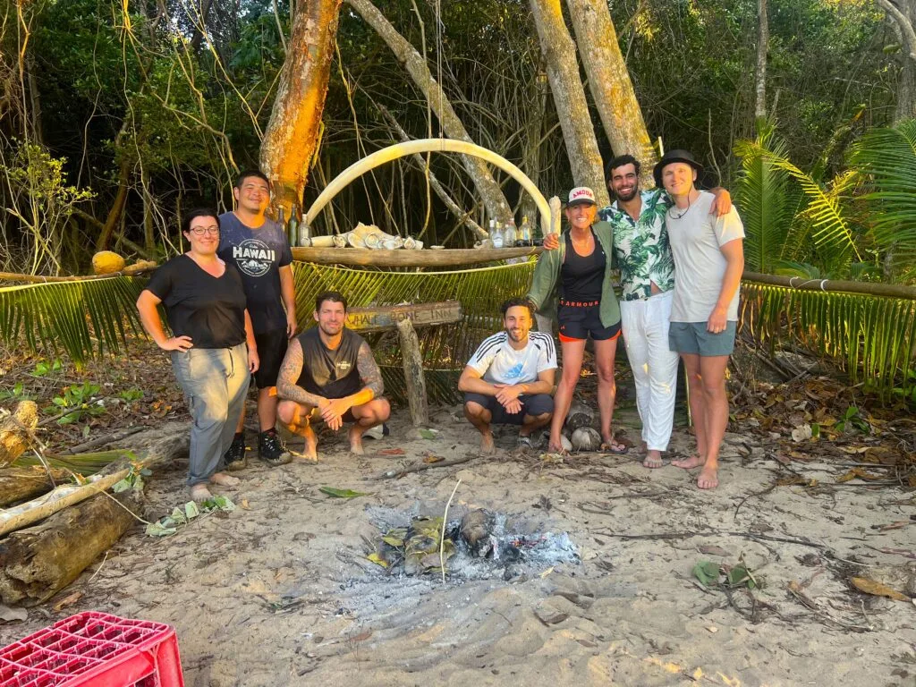 Seven people stand and kneel on a sandy beach in front of a campfire, surrounded by trees and greenery. A makeshift table with bottles and items is behind them, lit by the setting sun—a perfect scene for team building.