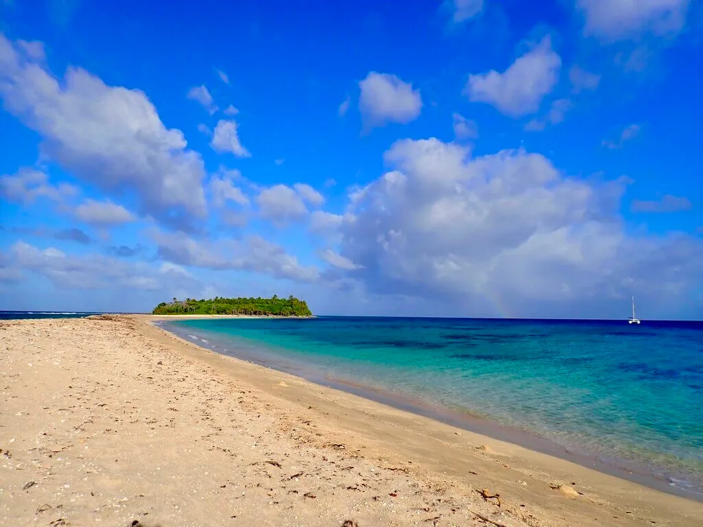Tonga in May: Tropical beach with turquoise water, white sand, and a small island under a blue sky with clouds.