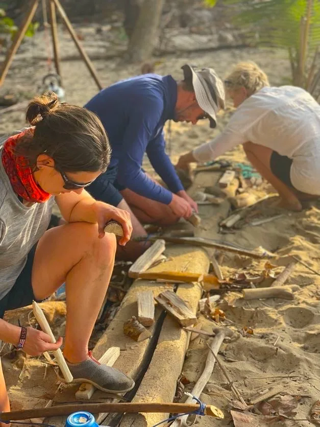 Three people sit on sand outdoors, focused on carving or shaping pieces of wood with hand tools. Sunlight illuminates their team building activity, surrounded by driftwood and natural debris, with trees visible in the background.