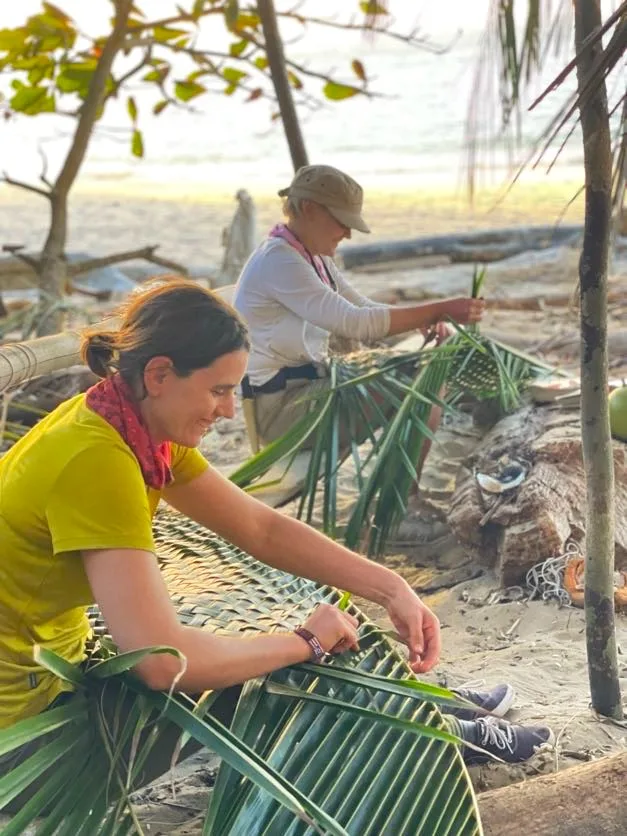 Team building activity: Two women weaving palm leaves on a beach.