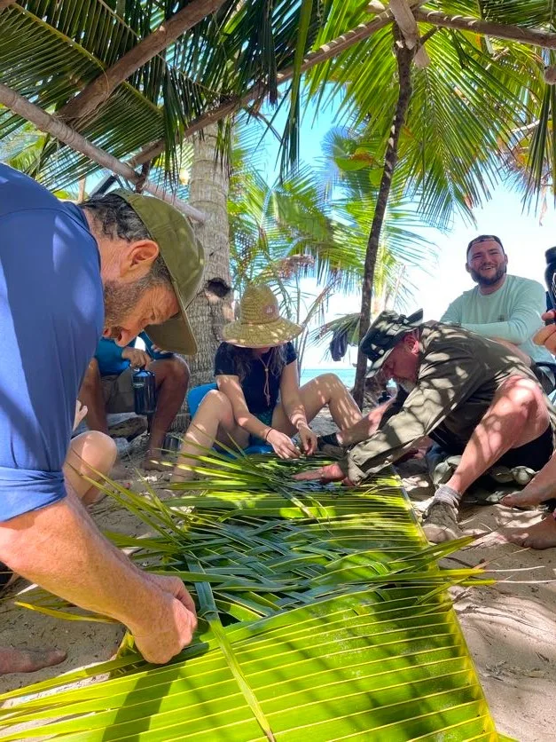 A group of people sit under palm trees on a sunny beach, weaving palm fronds together as a team building activity. Some focus on their hands while others smile and watch, enjoying the collaborative outdoor experience.