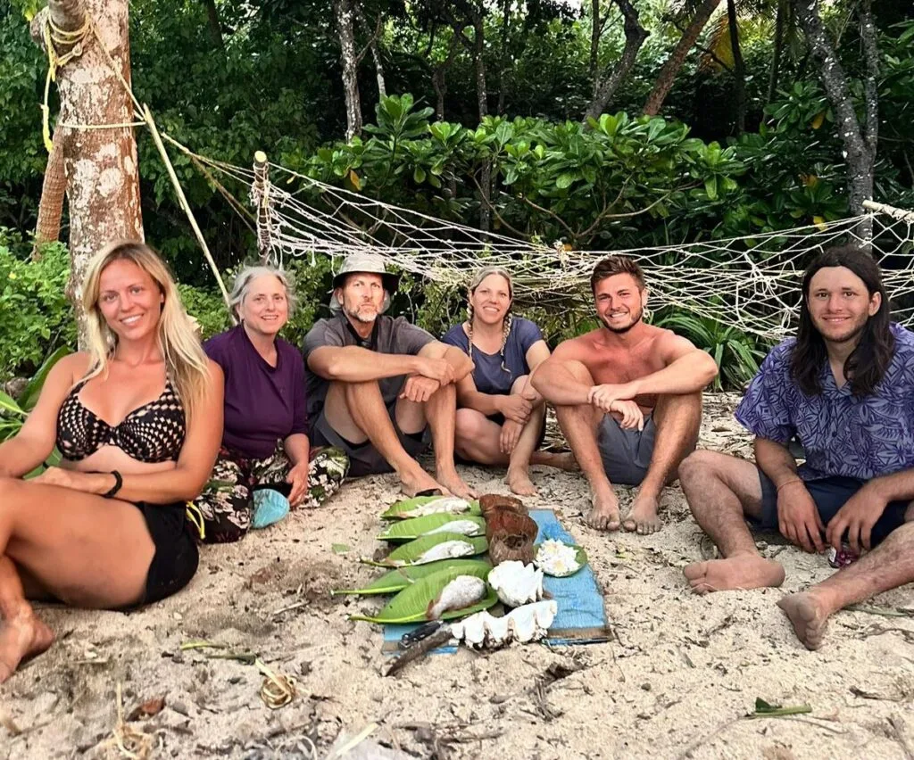 Six people sit barefoot on sandy ground near a hammock and lush greenery, sharing food wrapped in leaves on a blue mat—a relaxed outdoor team building gathering in a natural setting.