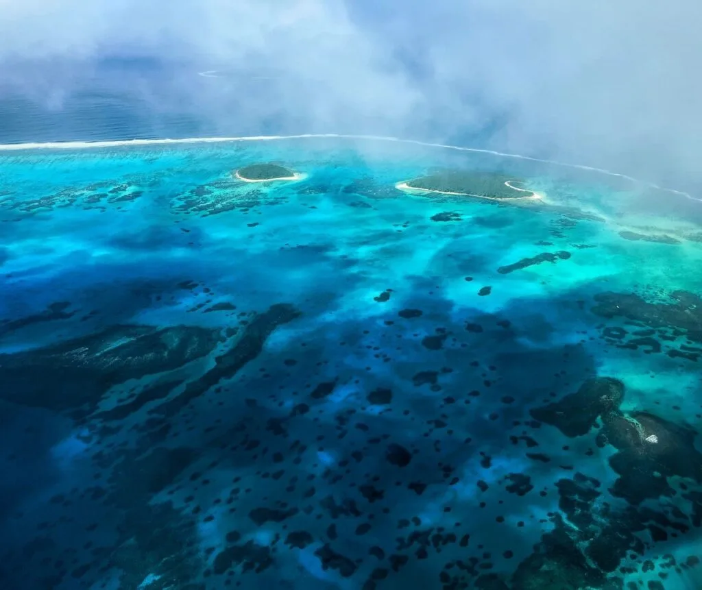 Aerial view of a vibrant turquoise ocean with scattered dark patches of coral reefs—perfect for team building—plus small sandy islands, partly covered by light mist or clouds near the horizon.