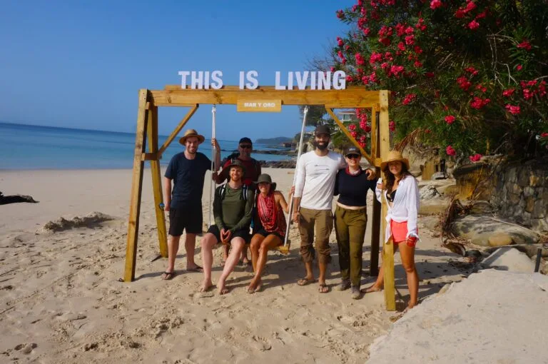 Seven people pose and smile on a sandy beach under a wooden swing frame that reads THIS IS LIVING, celebrating a corporate reward. The ocean and pink flowering bushes shine in the background beneath the clear blue sky.