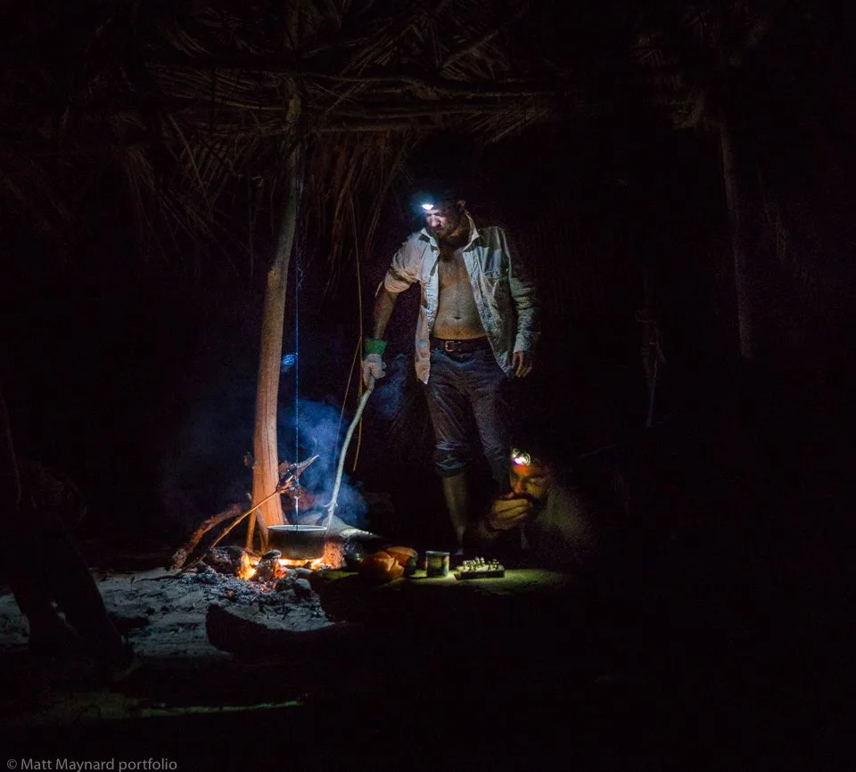 Team building: Two men with headlamps cooking over a fire in a dark hut.
