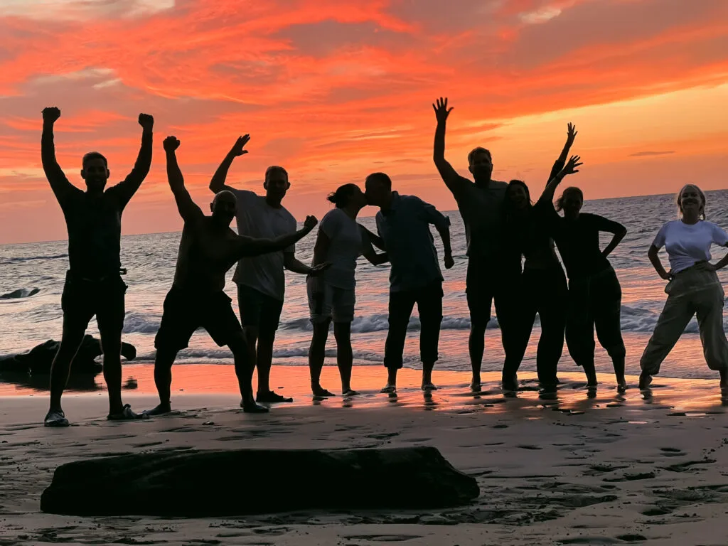 A group of friends are celebrating the sunset on the beach. Silhouetted against the vibrant orange and pink sky, they are posing and enjoying the beautiful moment together. The image captures a sense of joy and camaraderie as they share this special time.
