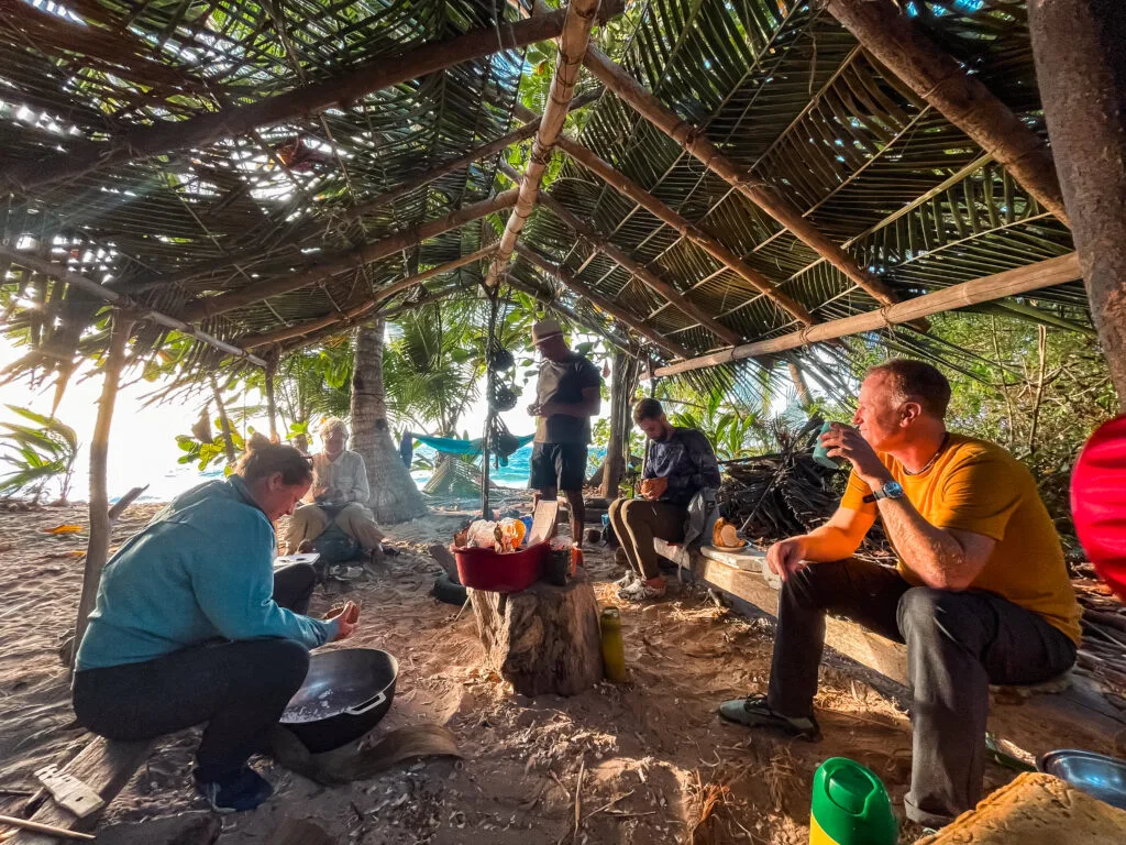 A group of people are enjoying a meal under a palm frond-thatched shelter on a beach. Several individuals, likely researchers or travelers, are preparing and eating a meal around a small fire. The scene suggests a relaxed, informal gathering, perhaps a break from fieldwork or exploration. The natural setting and the simple shelter highlight a connection with nature.