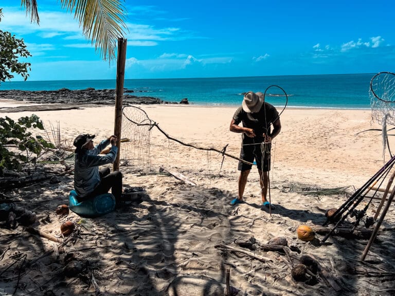 A Day in the Life of a Desert Island Survival Adventurer 10 Two individuals are setting up fishing nets on a sandy beach. One person, seated, is attaching the net to a wooden pole, while the other, standing, is working on securing the net's supporting lines. The scene suggests a traditional fishing method, utilizing natural materials and techniques. The bright blue sky and ocean in the background highlight the idyllic coastal location.