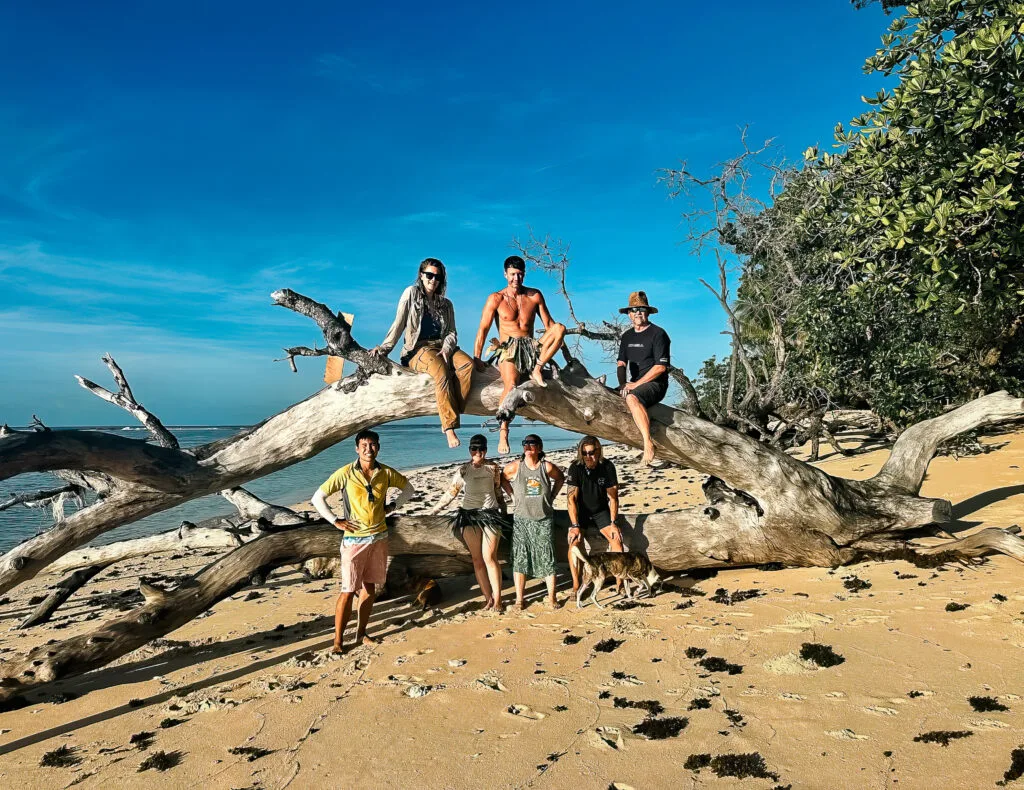 Group photo on a beach with a large driftwood log, evoking the Robinson Crusoe castaway setting.