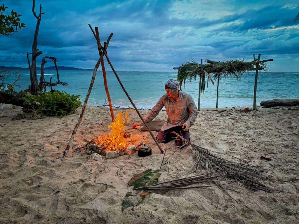 Robinson Crusoe-style survival: Man tending a beach campfire under a makeshift shelter at dusk.