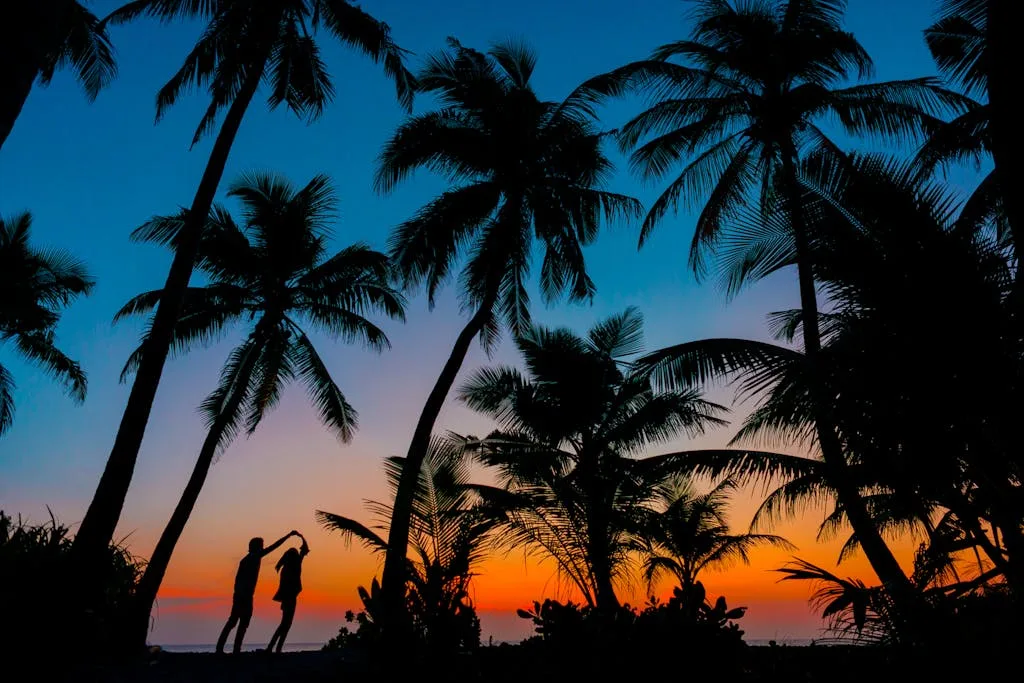 Silhouetted couple dancing under palm trees at tropical beach sunset, capturing romance and idyllic scenery.