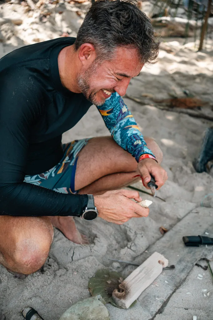 Man smiling while using a knife to carve wood for a bow drill fire starter on a beach.