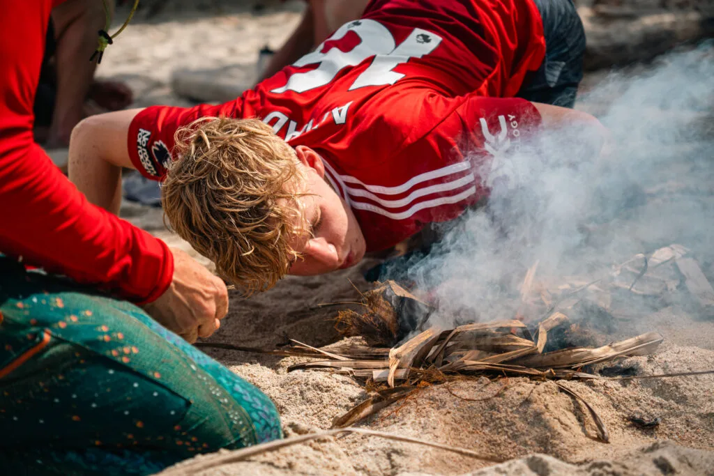 Man using a bow drill to start a fire on a sandy beach, smoke rising.