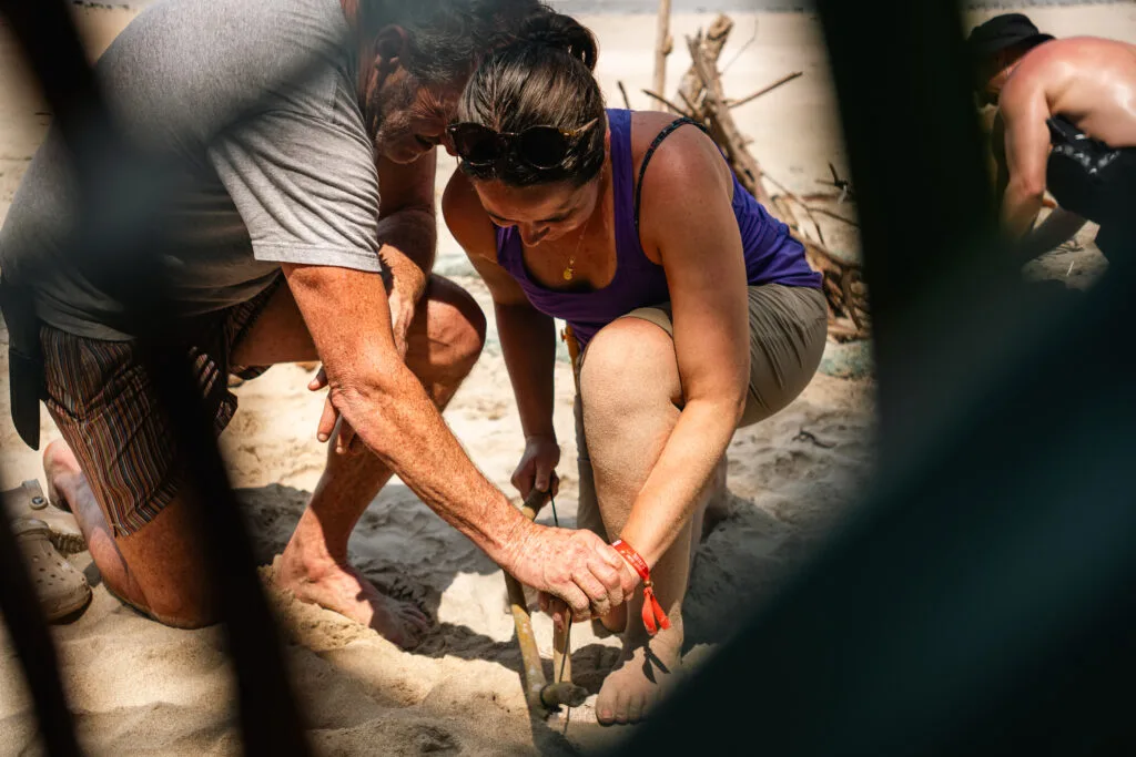 Man and woman using a bow drill to start a fire on a sandy beach, part of a survival skills demonstration.