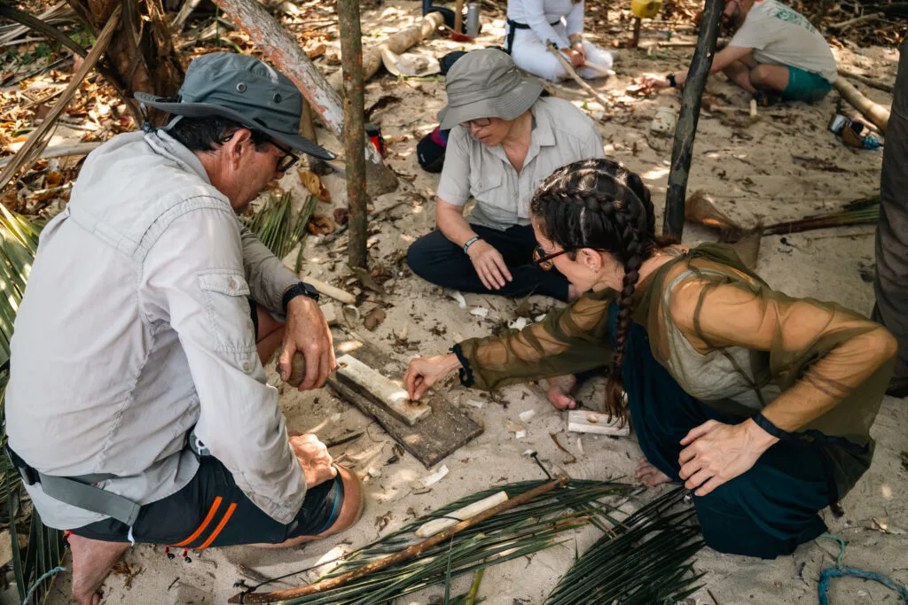 People learning how to start a fire with a bow drill on a sandy beach, survival skills
