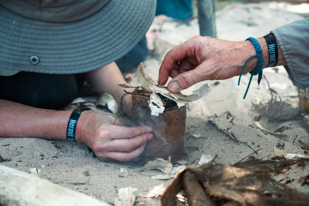 Starting a fire with a bow drill. Hands tending to the tinder bundle with smoke rising.