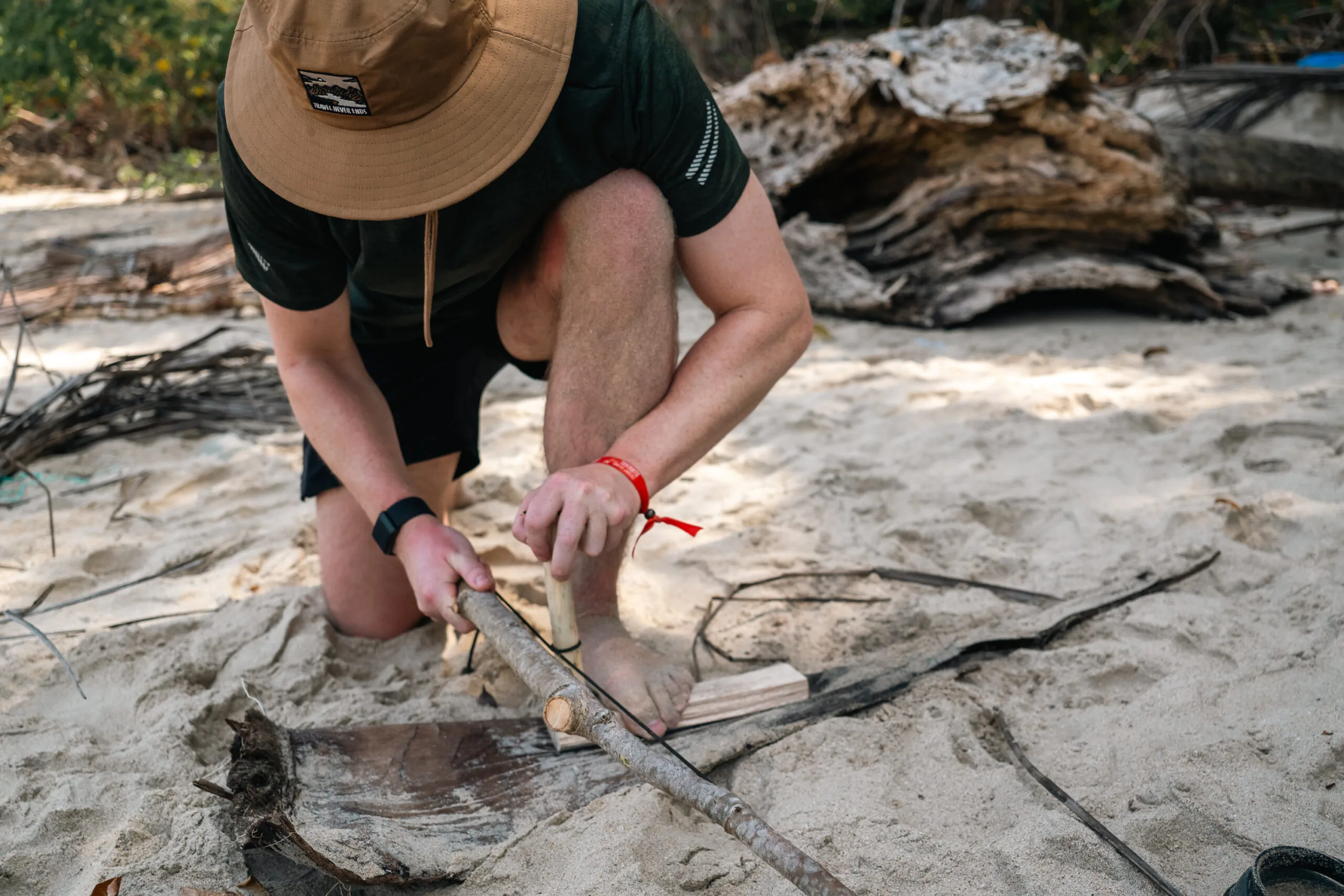 Man using a bow drill to start a fire on a sandy beach, wearing a hat.