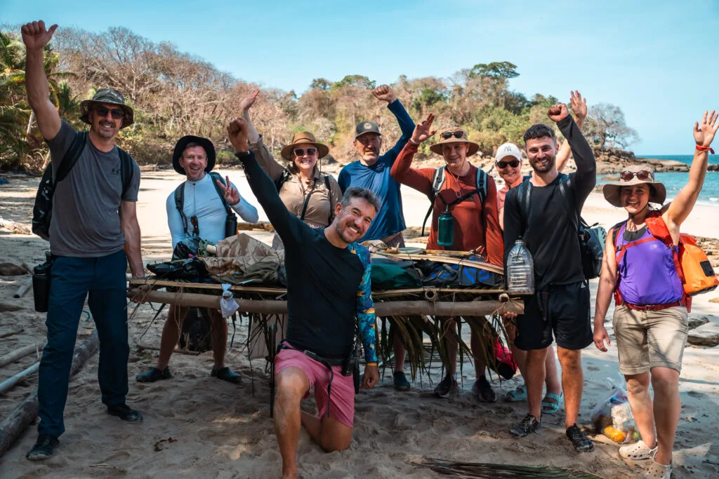 Group of people posing with a makeshift raft on a sandy beach, possibly related to famous castaways.