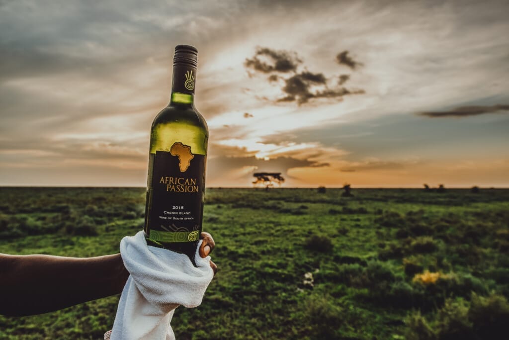 Hadza Tribal Bushcraft Immersion & Luxury Safari 10 A hand holding a bottle of African Passion wine wrapped in a white cloth, set against a grassy plain and dramatic sunset—an experience reminiscent of Hadza Tribal Bushcraft Immersion & Luxury Safari adventures.