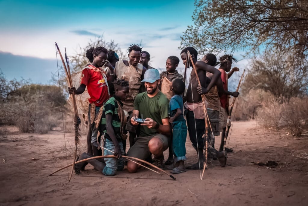 The Last Hunter-Gatherers: Inside the Ancient World of Tanzania’s Hadza Tribe 7 During a Hadza Tribal Bushcraft Immersion & Luxury Safari, a group stands on sandy ground outdoors—most holding bows—while one person kneels to show something on a device to several children, with trees and shrubs visible in the background.
