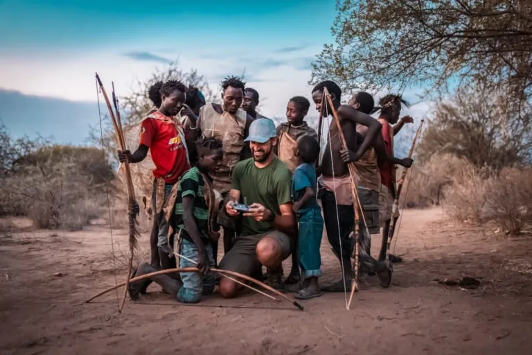 During a Hadza Tribal Bushcraft Immersion & Luxury Safari, a group stands on sandy ground outdoors—most holding bows—while one person kneels to show something on a device to several children, with trees and shrubs visible in the background.