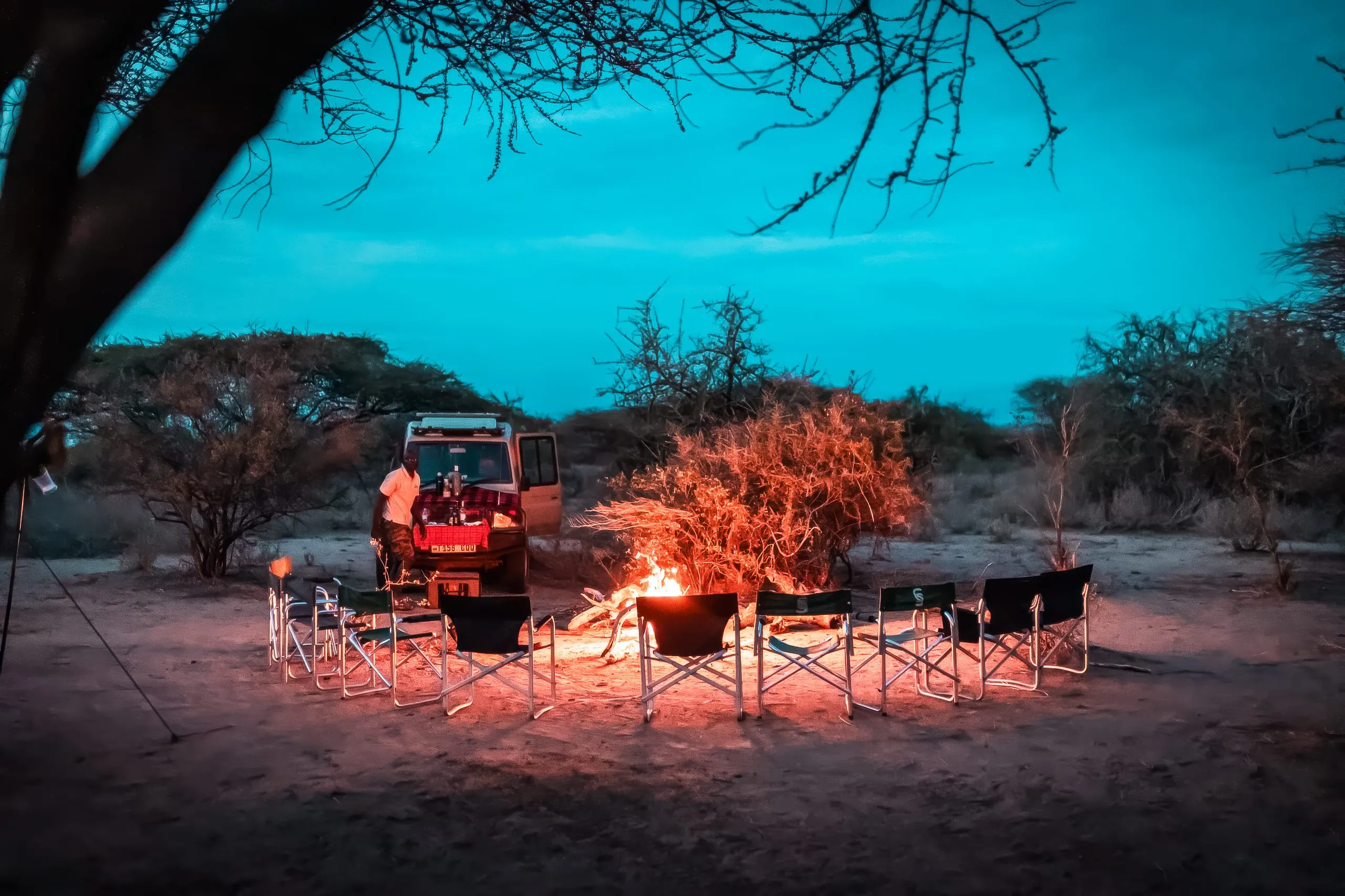 A circle of empty camping chairs surrounds a campfire near a vehicle at dusk in a dry, wooded area, an inviting scene from the Hadza Tribal Bushcraft Experience & Luxury Safari. A person stands nearby under the tranquil blue evening sky.