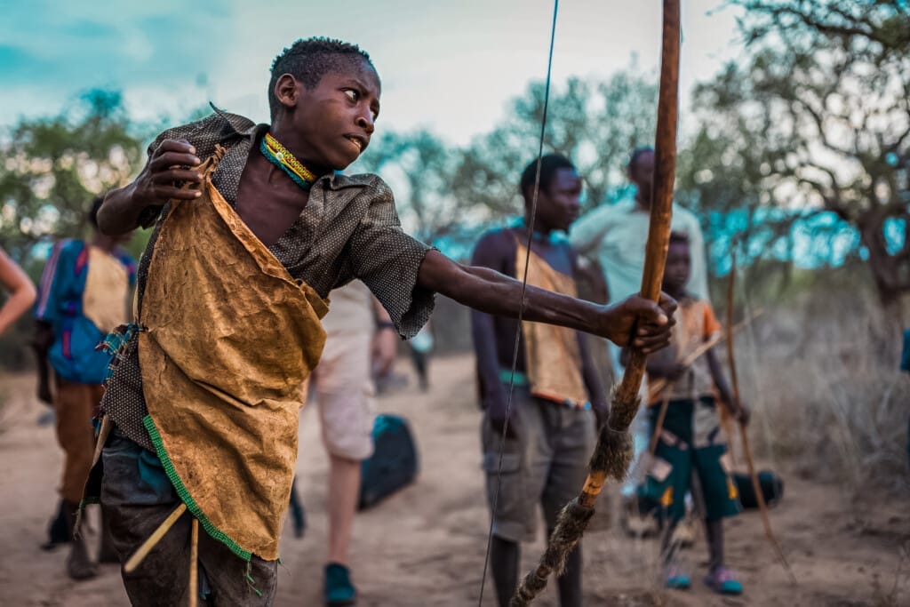 The Last Hunter-Gatherers: Inside the Ancient World of Tanzania’s Hadza Tribe 1 A boy in traditional clothing aims a bow and arrow outdoors during the Hadza Tribal Bushcraft Immersion & Luxury Safari, surrounded by children and adults in a scenic rural setting.