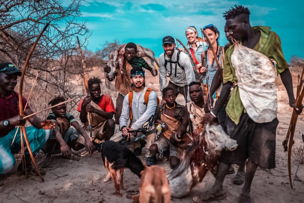 The Last Hunter-Gatherers: Inside the Ancient World of Tanzania’s Hadza Tribe 4 A group of people, including locals and tourists with backpacks, pose outdoors beside a freshly hunted animal and two dogs in a dry, rural landscape during a Hadza Tribal Bushcraft Immersion & Luxury Safari.