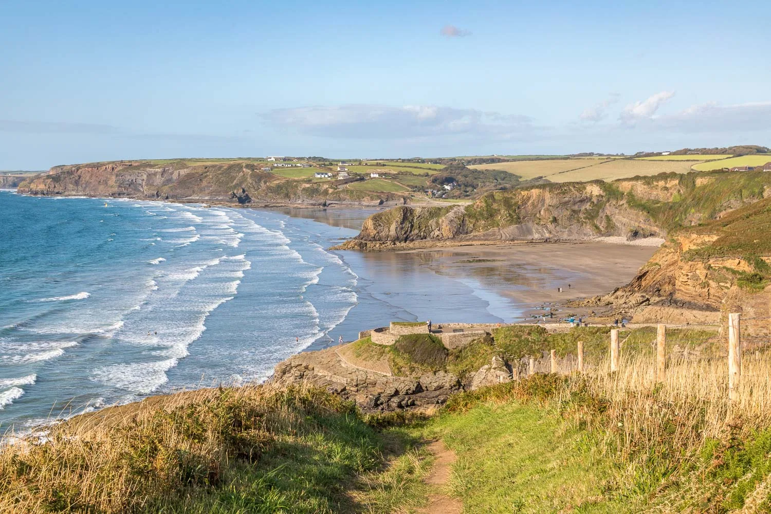 Pembrokeshire coastline view with sandy beach, cliffs, and rolling green hills under a blue sky.