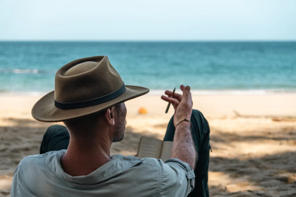 Man in hat relaxes on beach, writing in notebook. Enjoying a day without technology.