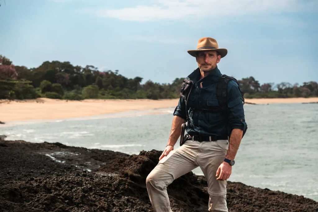 Man in safari attire standing on rocks near the ocean, ready to travel light.