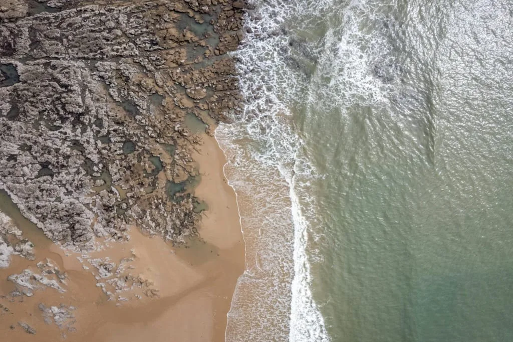A stunning aerial view highlighting the meeting of rocky terrain and ocean waves along a sandy shore.