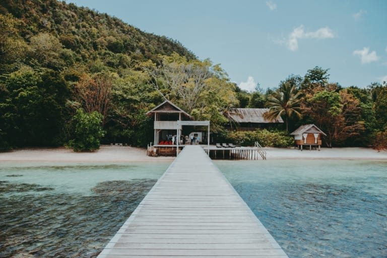 Tropical beach resort with a long wooden pier leading to bungalows and lush green hills.