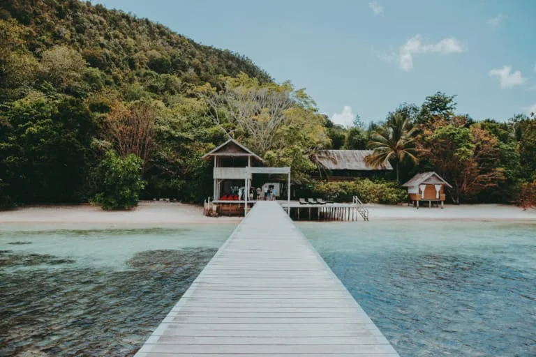 Tropical beach resort with a long wooden pier leading to bungalows and lush green hills.